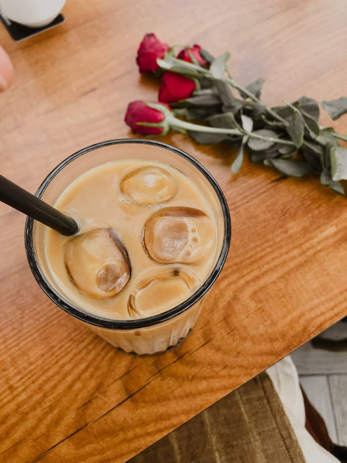 A glass of iced coffee with a black straw on a wooden table beside red roses. Perfect for a refreshing break.